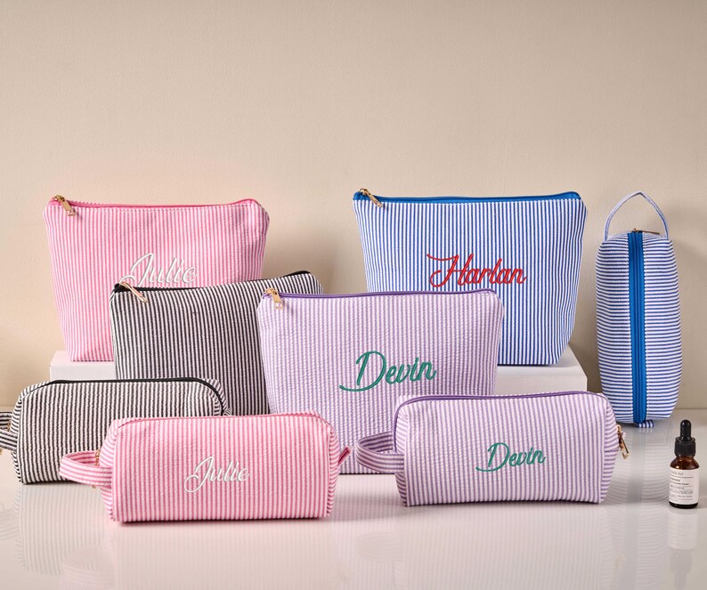 Pode incluir: Cole&ccedil;&atilde;o de n&eacute;cessaires personalizados em v&aacute;rios tamanhos e cores, incluindo rosa, azul, preto e lavanda. Cada bolsa apresenta um padr&atilde;o de listras seersucker e nomes bordados em escrita cursiva. Z&iacute;peres dourados e uma pequena al&ccedil;a vis&iacute;veis.