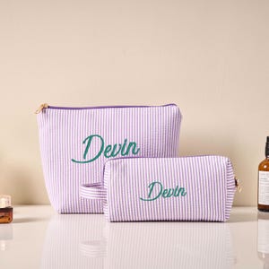 Pode incluir: Duas bolsas de cosm&eacute;ticos listradas em lavanda e branco. A bolsa maior tem uma al&ccedil;a e ambas as bolsas t&ecirc;m o nome "Devin" bordado em verde. Ambas as bolsas t&ecirc;m z&iacute;peres dourados.