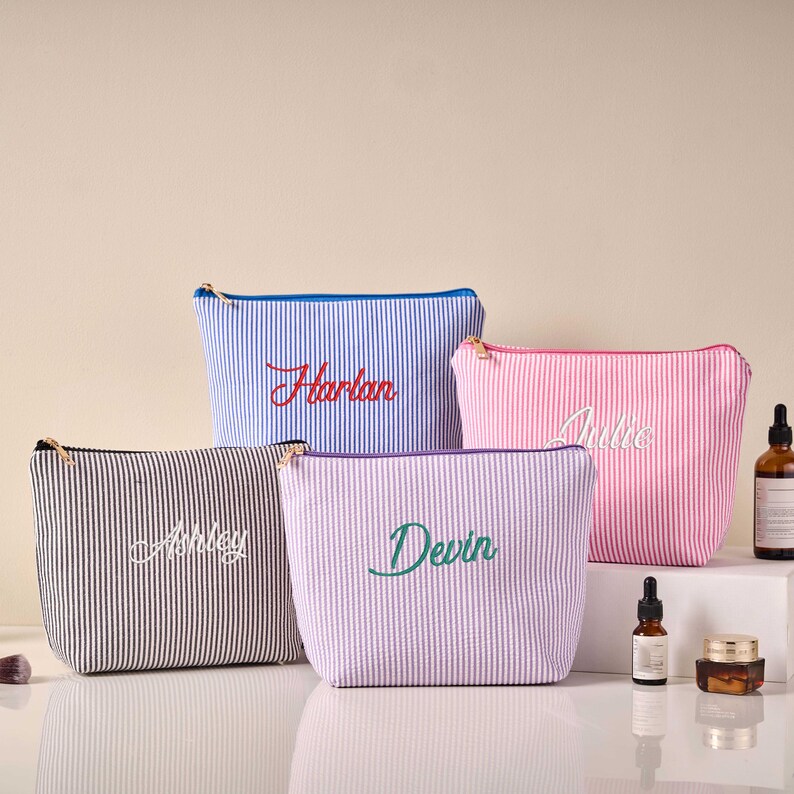 Pode incluir: Quatro bolsas de cosm&eacute;ticos listradas em azul, rosa, preto e lavanda. Cada bolsa tem um nome diferente bordado: Harlan, Julie, Ashley e Devin. As bolsas t&ecirc;m fecho de z&iacute;per e est&atilde;o sobre uma superf&iacute;cie branca.