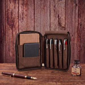 May include: A brown leather zippered organizer case featuring an interior pocket holding a tan notebook and a black notebook, alongside five elastic pen loops containing various writing instruments. A fountain pen lies nearby.
