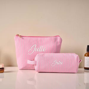 Pode incluir: Duas bolsas de cosm&eacute;ticos listradas em rosa e branco, uma maior e outra menor, com o nome "Julie" bordado em branco. A bolsa maior tem um z&iacute;per dourado e a bolsa menor tem uma al&ccedil;a.