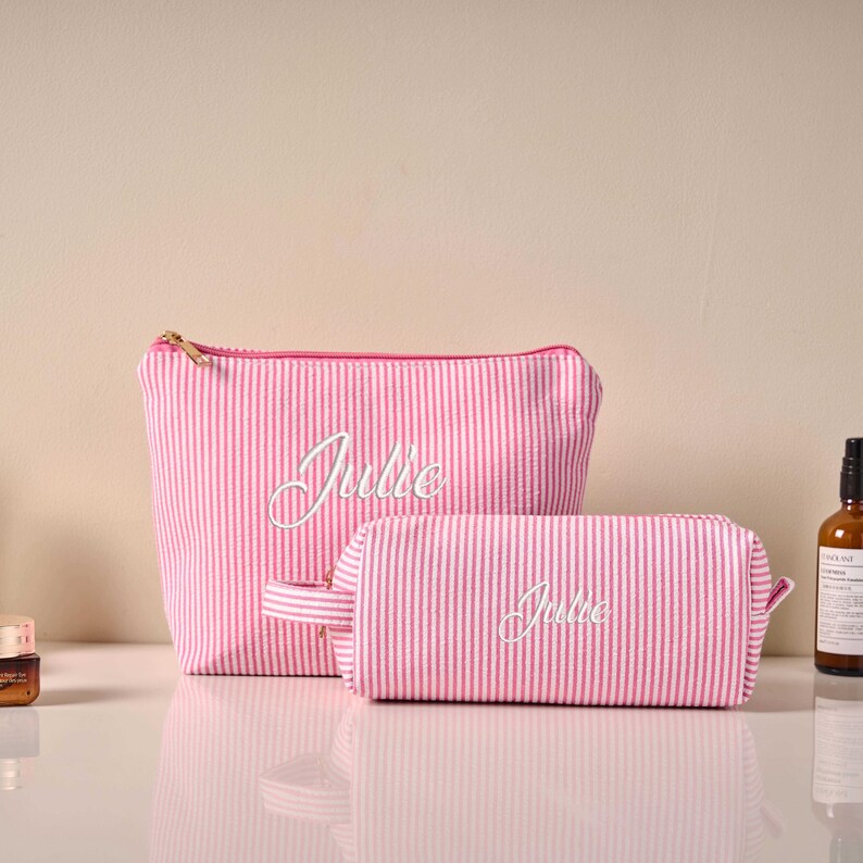 Pode incluir: Duas bolsas de cosm&eacute;ticos listradas em rosa e branco, uma maior e outra menor, com o nome "Julie" bordado em branco. A bolsa maior tem um z&iacute;per dourado e a bolsa menor tem uma al&ccedil;a.