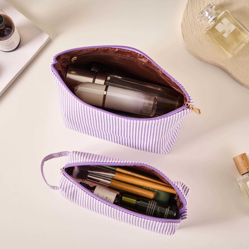 Pode incluir: Duas bolsas de cosm&eacute;ticos abertas e a condizer, com riscas verticais roxas e brancas. A bolsa maior cont&eacute;m v&aacute;rias garrafas e tubos. A bolsa menor cont&eacute;m pinc&eacute;is de maquilhagem e outros produtos de beleza. Ambas as bolsas t&ecirc;m interiores roxos e fechos dourados.