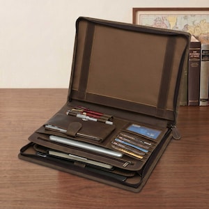 May include: Open brown leather portfolio with multiple compartments. Features include pen holders, card slots, and a tablet sleeve. The portfolio is on a wooden surface, with pens, a tablet, and cards visible.