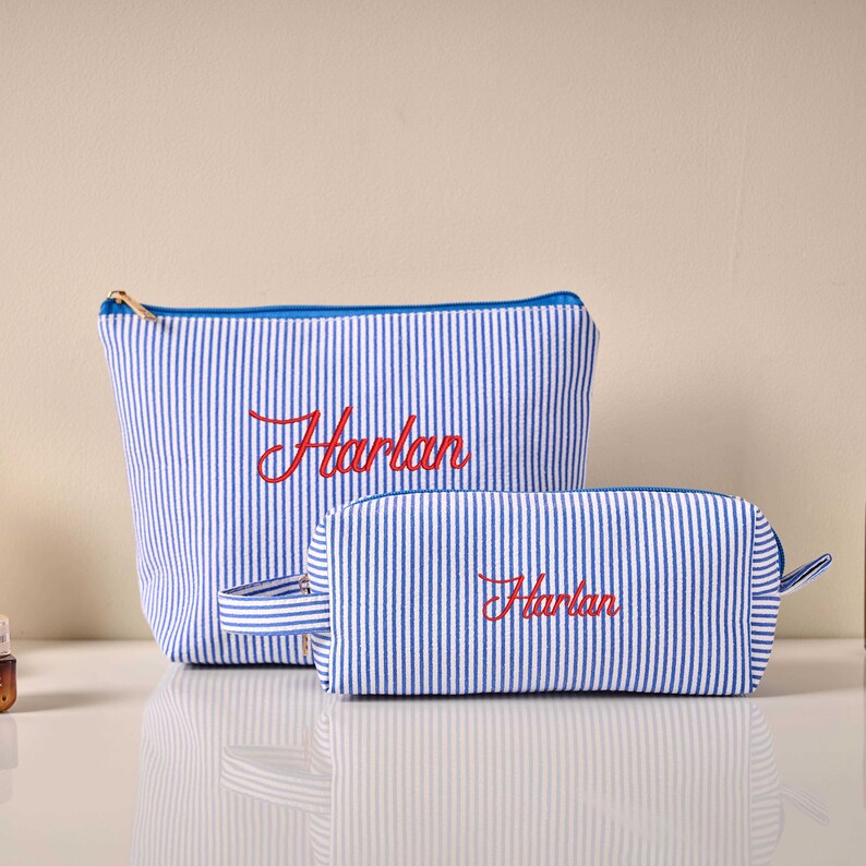Pode incluir: Duas bolsas de cosm&eacute;ticos a condizer com riscas verticais azuis e brancas. A bolsa maior tem um fecho azul e o nome "Harlan" bordado a vermelho. A bolsa menor tem um fecho azul e o nome "Harlan" bordado a vermelho.