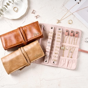 Könnte beinhalten: Ein hellbraunes und beigefarbenes Leder-Schmuck-Etui mit den Namen "Lisa" und "Daisy" auf der Vorderseite. Die Rolle ist geöffnet und zeigt ein rosafarbenes Innere mit Ringhaltern und Halskettenhaken. Verschiedene Ringe und Halsketten sind ausgestellt.