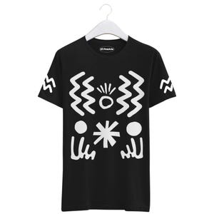 Peut inclure: T-shirt noir avec un motif abstrait blanc. Le motif comprend des lignes en zigzag, un cercle avec des lignes rayonnantes, une étoile et deux cercles avec des lignes en dessous. Le texte "El Fenicío" est sur le col.