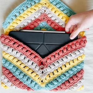 Puede incluir: Una funda de tableta de ganchillo hecha a mano con un patrón de arcoíris. La funda está hecha con una variedad de colores de hilo, incluyendo rosa, azul, amarillo y gris. Una tableta negra está dentro. La funda se cierra con un botón.