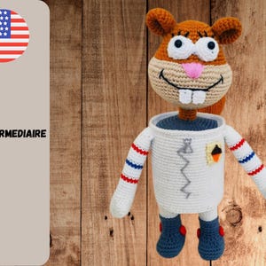 May include: Crocheted Sandy Cheeks doll in a white astronaut suit with blue boots and red and blue striped arms. The doll has a tan face with white teeth, a pink nose, and large white eyes. The background is a wooden surface.