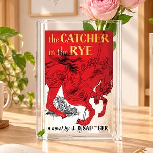 May include: A clear rectangular glass vase featuring a book cover insert for The Catcher in the Rye by J.D. Salinger, which displays a red carousel horse illustration. Two light pink roses are placed inside the vase, extending from the top.