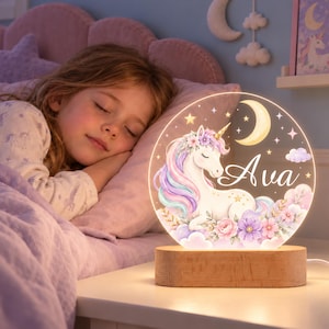 Puede incluir: Una luz nocturna circular de acrílico con un unicornio blanco de crines arcoíris pastel, detalles florales, una luna creciente y estrellas. El nombre Ava está impreso en letra blanca. La luz se apoya sobre una base de madera rectangular.