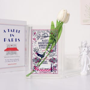 May include: A clear glass vase with a white tulip and green leaves. The vase displays the cover of "Alice's Adventures in Wonderland & Other Stories" by Lewis Carroll. A book titled "A Table in Paris" is visible in the background.