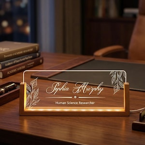 May include: A personalized desk nameplate with the name "Sophia Murphy" and the title "Human Science Researcher" is illuminated by LED lights. The nameplate is set in a wooden frame, with a leaf design. The desk is made of wood.