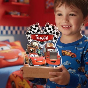 Può includere: Una luce notturna in legno con personaggi del film Cars, tra cui Saetta McQueen, Cricchetto e Mack. La luce notturna ha un design a bandiera a scacchi e il nome "Rowan" sopra. Il bambino indossa un pigiama blu a tema Cars.