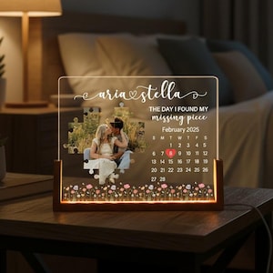 May include: A personalized acrylic night light with a wooden base. The light features a photo within a puzzle design, the names "Aria & Stella", and a calendar highlighting February 2025. The base is illuminated with a warm glow, and the bottom has a floral design.