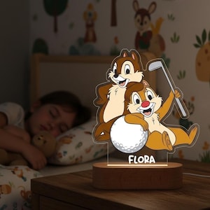 Op de afbeelding: Een nachtlampje met Chip en Dale met een golfbal en club. De basis is van hout en de naam "FLORA" staat op het licht. Op de achtergrond slaapt een kind in een bed met een patroon op het dekbed.