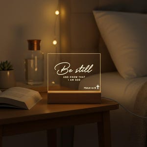 May include: A lighted square acrylic night light with the words "Be still and know that I am God" in white script. The night light sits on a wooden base and is on a wooden nightstand. A book, a small plant, and a glass bottle are also on the nightstand.