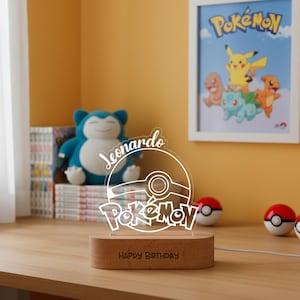 Puede incluir: Luz de noche de Pokémon de acrílico transparente, con el logotipo de Pokémon, un diseño de Poké Ball y el nombre "Leonardo". La base de madera dice "HAPPY BIRTHAY". Un póster de Pokémon enmarcado y un peluche están en el fondo.