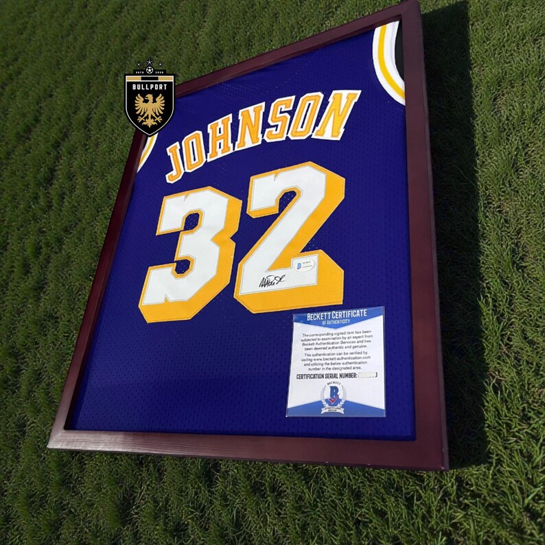 Puede incluir: Camiseta azul enmarcada con el nombre "JOHNSON" y el n&uacute;mero "32" en amarillo y blanco. La camiseta se muestra en un marco marr&oacute;n oscuro, con un Certificado de Autenticidad Beckett.