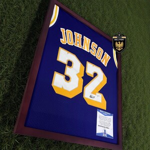 Puede incluir: Camiseta de baloncesto azul enmarcada con el nombre "JOHNSON" y el n&uacute;mero "32" en amarillo y blanco. La camiseta se muestra en un marco marr&oacute;n oscuro, con un certificado de autenticidad visible.