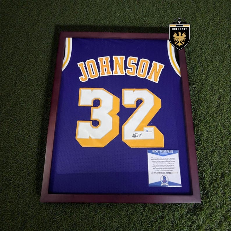Puede incluir: Camiseta de baloncesto morada enmarcada con letras y n&uacute;meros dorados. La camiseta muestra el nombre "JOHNSON" y el n&uacute;mero "32". Se incluye un certificado de autenticidad Beckett. El marco es de color marr&oacute;n oscuro.