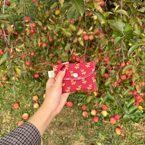 May include: A small, red fabric pouch with a yellow floral pattern and a white button closure. The pouch is held in front of a background of apple trees with red apples on the branches and on the ground.