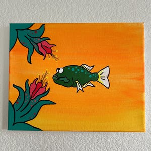 May include: A whimsical painting on canvas featuring a green fish with white fins and yellow accents against an orange background. Two stylized flowers with red petals and green leaves frame the fish. The artwork is mounted on a white wall.