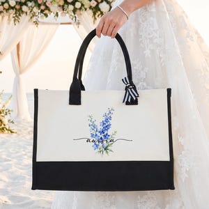 May include: A rectangular canvas tote bag with a cream-colored upper section and a solid black base. The front features a watercolor illustration of blue delphinium flowers with the name aurora written in black script. It has black handles and a small black and white striped ribbon tied to one handle.