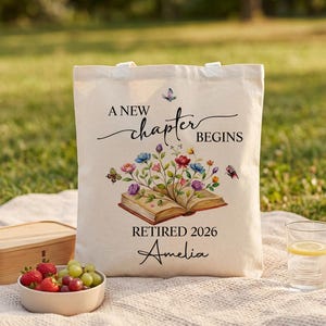 Peut inclure: Sac fourre-tout crème avec le texte "A New Chapter Begins" et "Retired 2026 Amelia". Le sac présente un livre ouvert avec des fleurs colorées et des papillons. Une boîte en bois, un bol de fruits et un verre d'eau sont au premier plan.