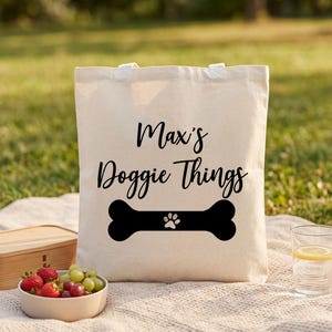 Puede incluir: Una bolsa de tela color crema con el texto negro impreso "Max's Doggie Things" sobre un gráfico de hueso de perro negro con una pequeña huella blanca en el centro. La bolsa tiene dos asas superiores y una forma rectangular.
