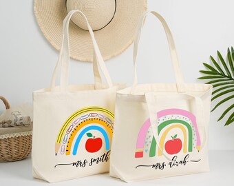 May include: Two cream tote bags with rainbow designs and the names "Mrs Smith" and "Mrs Sarah" in cursive. The rainbows feature pencils, rulers, and apples. A straw hat hangs in the background.