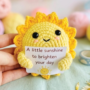May include: A crocheted yellow sun with a smiling face and black eyes holds a sign that reads "A little sunshine to brighten your day." The sun has a ruffled edge and is surrounded by colourful yarn balls and decorative tape.