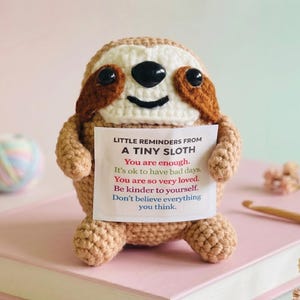 May include: A crocheted sloth doll with brown and beige yarn, holding a card with inspirational text. The sloth has black eyes and a smiling mouth. The card reads, "Little reminders from a tiny sloth."