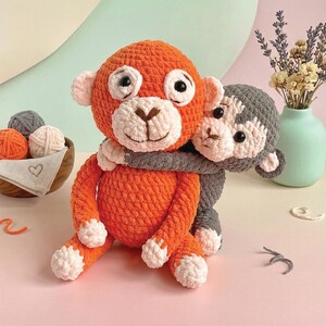 Op de afbeelding: Twee gehaakte apenknuffels, één oranje en één grijs, omarmen elkaar. De oranje aap heeft witte accenten op zijn gezicht, handen en voeten. Een houten kom met garenballen en een kleine vaas met bloemen staan op de achtergrond.