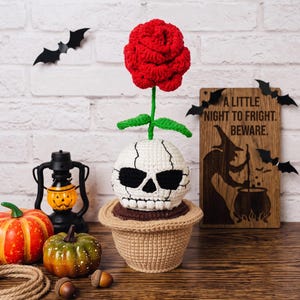 Könnte beinhalten: Ein gehäkelter Totenkopf in einem beigen Topf mit einer roten Rose, die oben herauswächst. Der Totenkopf ist weiß mit schwarzen Augenhöhlen und gestickten Details. Daneben stehen ein kleiner orangefarbener Kürbis, ein grüner Kürbis, eine schwarze Laterne und ein Holzschild.