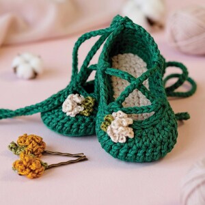 May include: A pair of dark green crocheted baby shoes with long laces and a cream-colored crocheted flower accent on the front of each shoe. Two gold-toned bobby pins with small yellow crocheted flower embellishments are placed in front of the shoes.