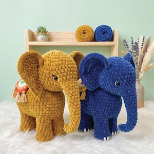 May include: Two handmade crochet elephant plush toys, one in mustard yellow and one in deep blue, both featuring large ears, dark eyes, and a small decorative blanket with tassels on their backs, crafted from soft, textured chenille yarn.