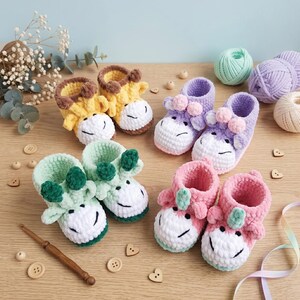 Può includere: Scarpine da neonato fatte a mano all'uncinetto in vari colori, tra cui giallo, verde, rosa e viola. Ogni paio presenta un design a forma di muso di animale con muso bianco e bocca sorridente. Esposte su una superficie di legno.