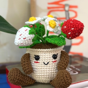 May include: A crocheted strawberry plant with a smiling face, black eyes, and brown arms and legs. The plant pot is beige, and the flowers are white with yellow centers. The strawberries are red and white.