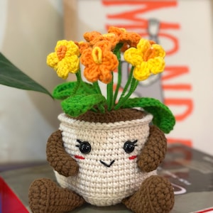 Puede incluir: Una maceta de ganchillo con una cara sonriente, brazos y piernas, que sostiene flores de ganchillo amarillas y naranjas. La maceta es de color blanquecino con detalles marrones. Las flores tienen tallos y hojas verdes.