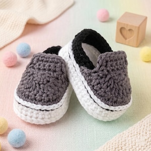 May include: A pair of handmade crochet baby shoes featuring a dark gray upper, a black collar, and a white sole. The shoes are crafted with a visible yarn texture and a slip-on design, mimicking the appearance of classic low-top sneakers.
