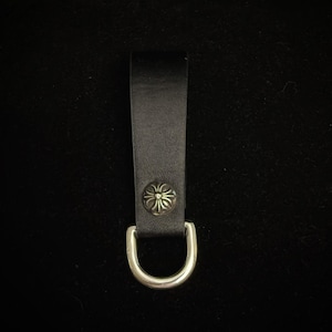 Puede incluir: Un llavero de cuero negro con una anilla en D de metal plateado y un botón decorativo plateado. El botón tiene un diseño floral. El llavero se muestra sobre un fondo negro.