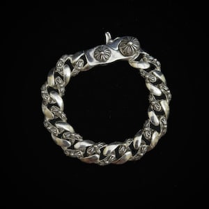 May include: A silver bracelet with a curb chain design. The bracelet has a decorative clasp with floral embellishments. The chain links are also detailed with similar floral patterns. The bracelet is set against a black background.
