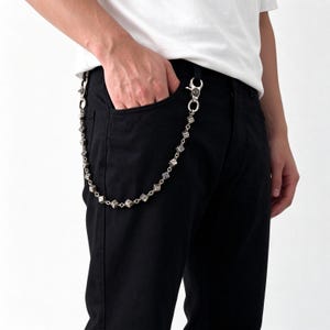 May include: A silver-toned metal wallet chain featuring a series of small, cube-shaped dice links connected by circular rings. The chain is attached to black trousers with two lobster claw clasps, each detailed with a circular emblem design.