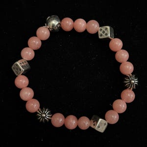 May include: A pink beaded bracelet with silver accents. The bracelet features round, light pink beads with silver dice-shaped beads and decorative silver charms. The bracelet is displayed against a black background.