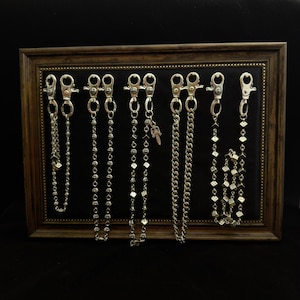 May include: A selection of silver-coloured wallet chains with different chain designs and decorative charms. The chains are displayed against a black backdrop within a framed presentation. The frame is a dark brown colour.