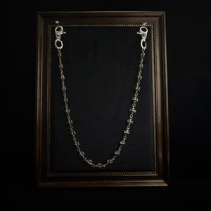 Pode incluir: Uma corrente prateada com um design floral, exibida sobre um fundo preto dentro de uma moldura. A corrente é fixada por dois fechos na parte superior. A moldura é castanha escura.