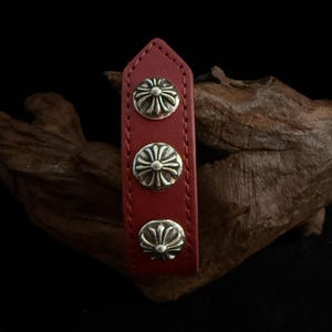May include: A red leather strap featuring three silver-coloured, decorative floral-style studs. The strap is rectangular with a pointed top. The studs are evenly spaced. The background is dark.