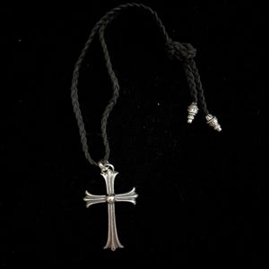Pode incluir: Colar com pingente de cruz prateada e cordão trançado preto. A cruz tem um design detalhado com bordas arredondadas e uma pequena conta no centro. O cordão é ajustável com contas decorativas prateadas nas extremidades.