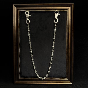 May include: A silver-toned wallet chain with a skull bead design. The chain has two clasp attachments and is displayed against a black background within a framed setting.